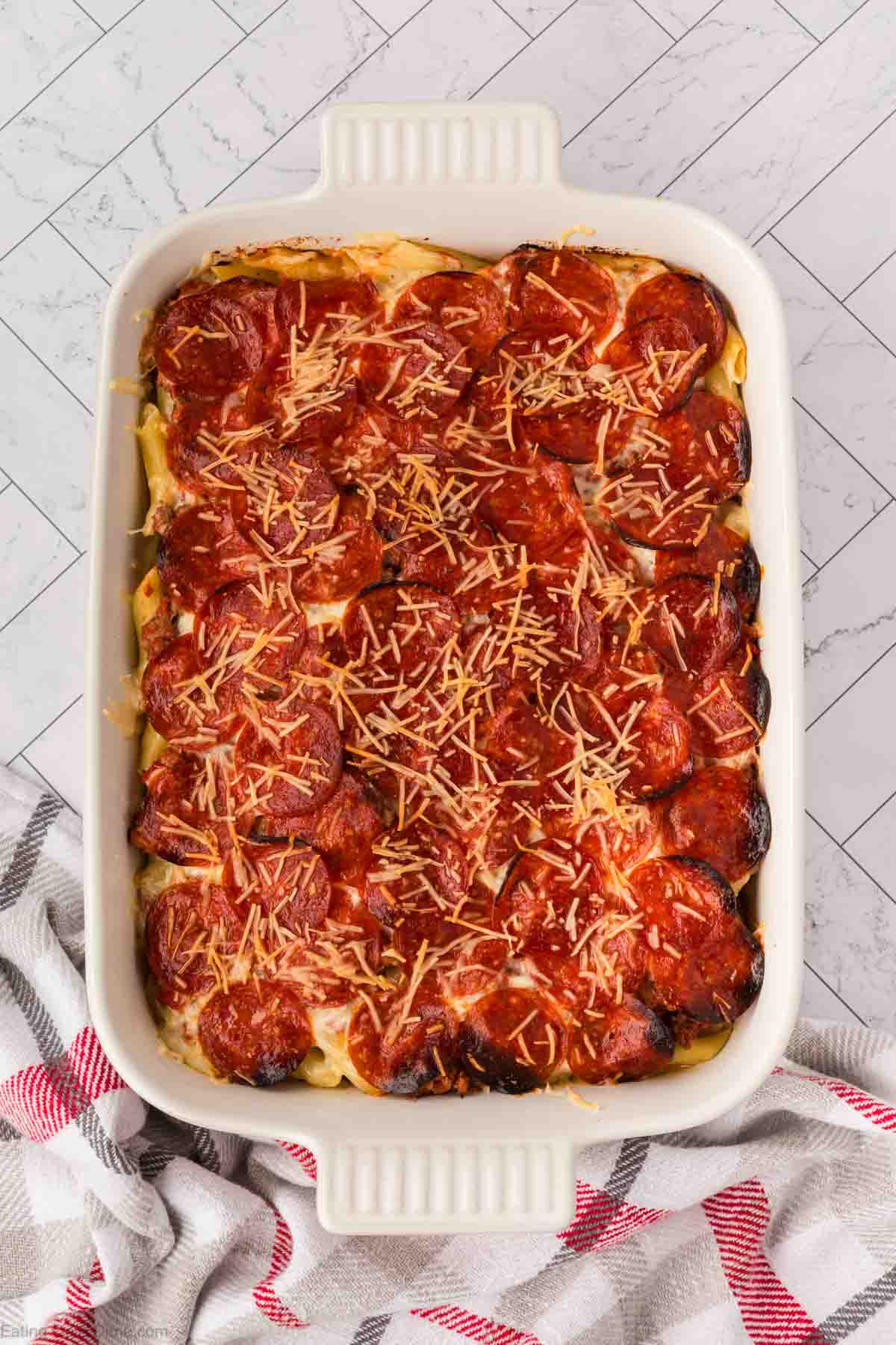 A rectangular baking dish filled with Pizza Casserole—baked pasta topped with slices of pepperoni and melted shredded cheese—sits on a marble surface next to a red and white kitchen towel.