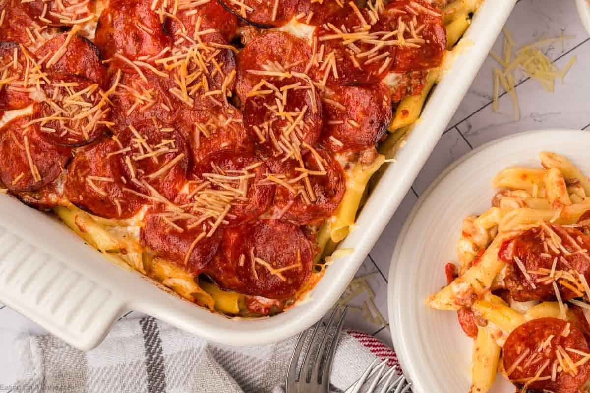 A Pizza Casserole, topped with pepperoni slices and melted cheese, sits baked in a white dish. A serving of the casserole rests on a nearby white plate, with a fork and napkin placed beside the dish.