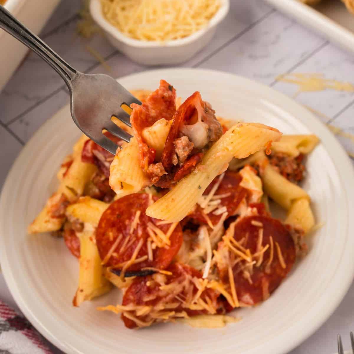 A fork holds a bite of Pizza Casserole, featuring baked penne pasta with melted cheese, pepperoni slices, and tomato sauce above a white plate of the same dish. A bowl of shredded cheese is visible in the background.