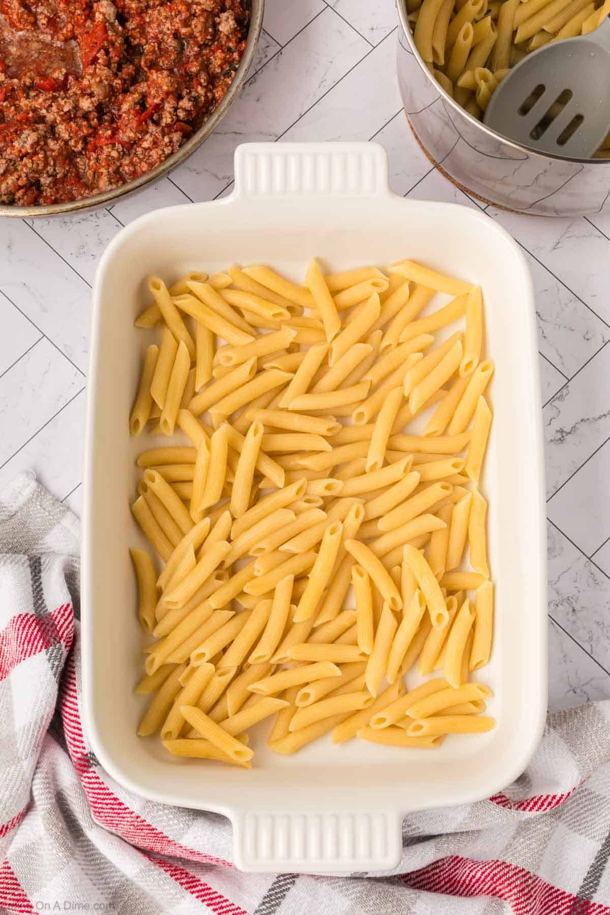 A white baking dish filled with uncooked penne pasta sits on a countertop, ready to become a delicious Pizza Casserole. Nearby, a pot of red meat sauce and a metal ladle await, with a red and white striped towel adding a cozy touch.