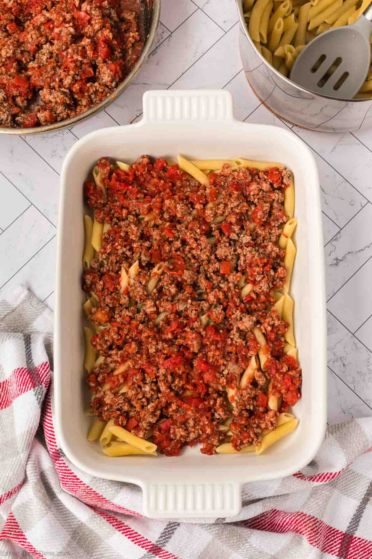 A white baking dish filled with penne pasta topped with ground beef and tomato sauce, resembling a Pizza Casserole. Next to it are a pan of sauce, a pot of cooked pasta, and a red and white towel partially visible on the left.