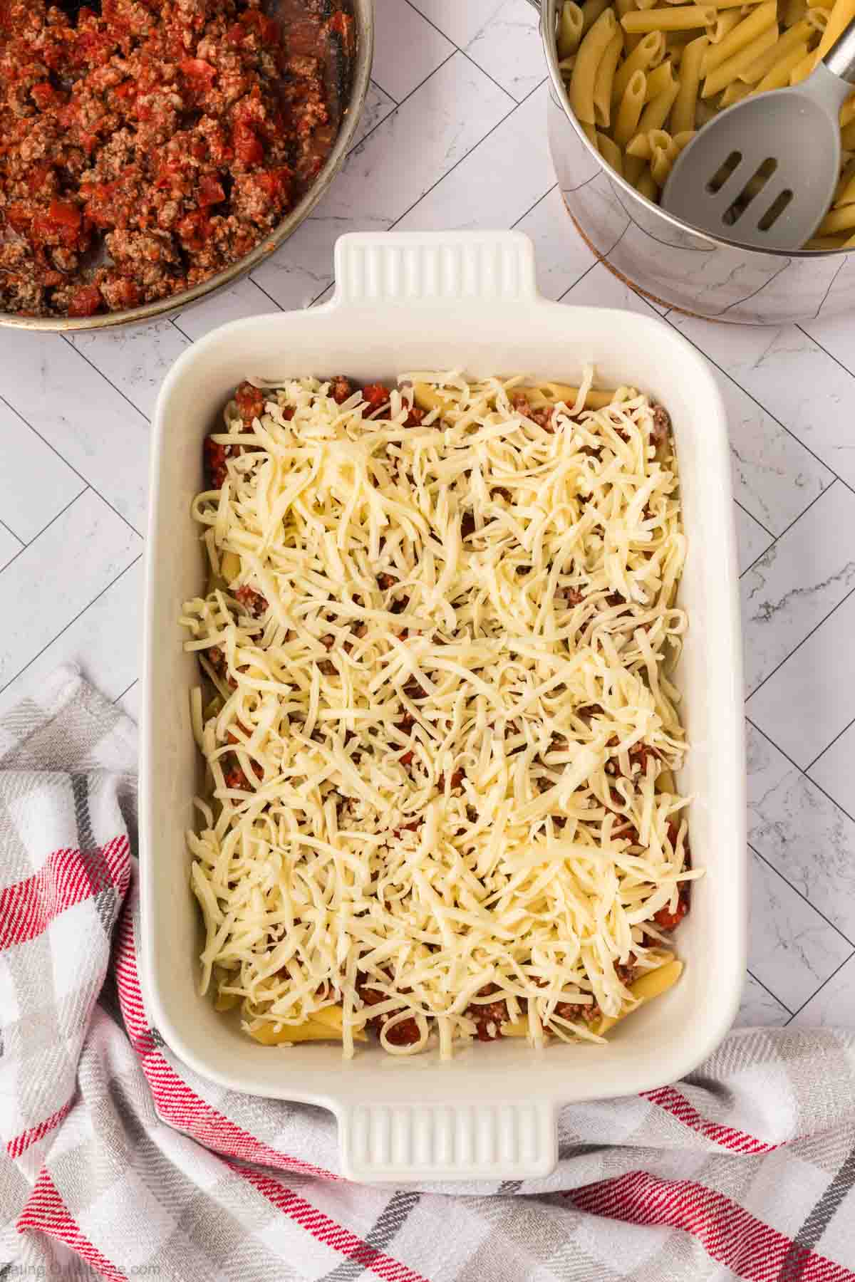A white baking dish filled with Pizza Casserole—pasta, meat sauce, and shredded cheese—sits on a countertop next to a bowl of meat sauce, a pot of cooked penne, and a red-and-white kitchen towel.