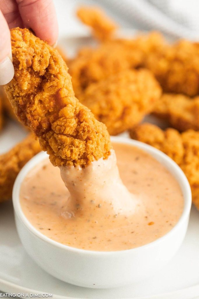 Close up image of chicken fingers and a bowl of chicken dipping sauce. Chicken finger being dipping into a bowl of sauce