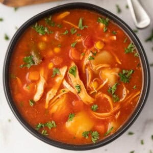 A bowl of crockpot chicken vegetable soup with shredded chicken, potatoes, carrots, peas, corn, and parsley garnish, served in a black bowl on a white surface.