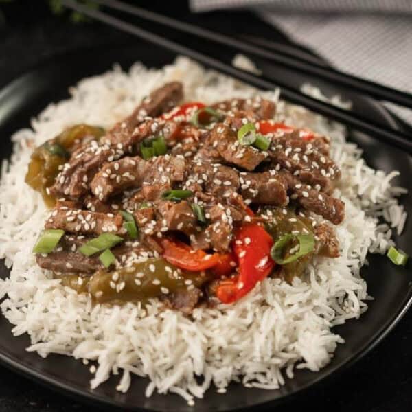 A plate of white rice topped with stir-fried beef strips, red and green bell peppers, sliced green onions, and sprinkled with sesame seeds—just like a delicious Crockpot Pepper Steak Recipe. Chopsticks rest in the background.