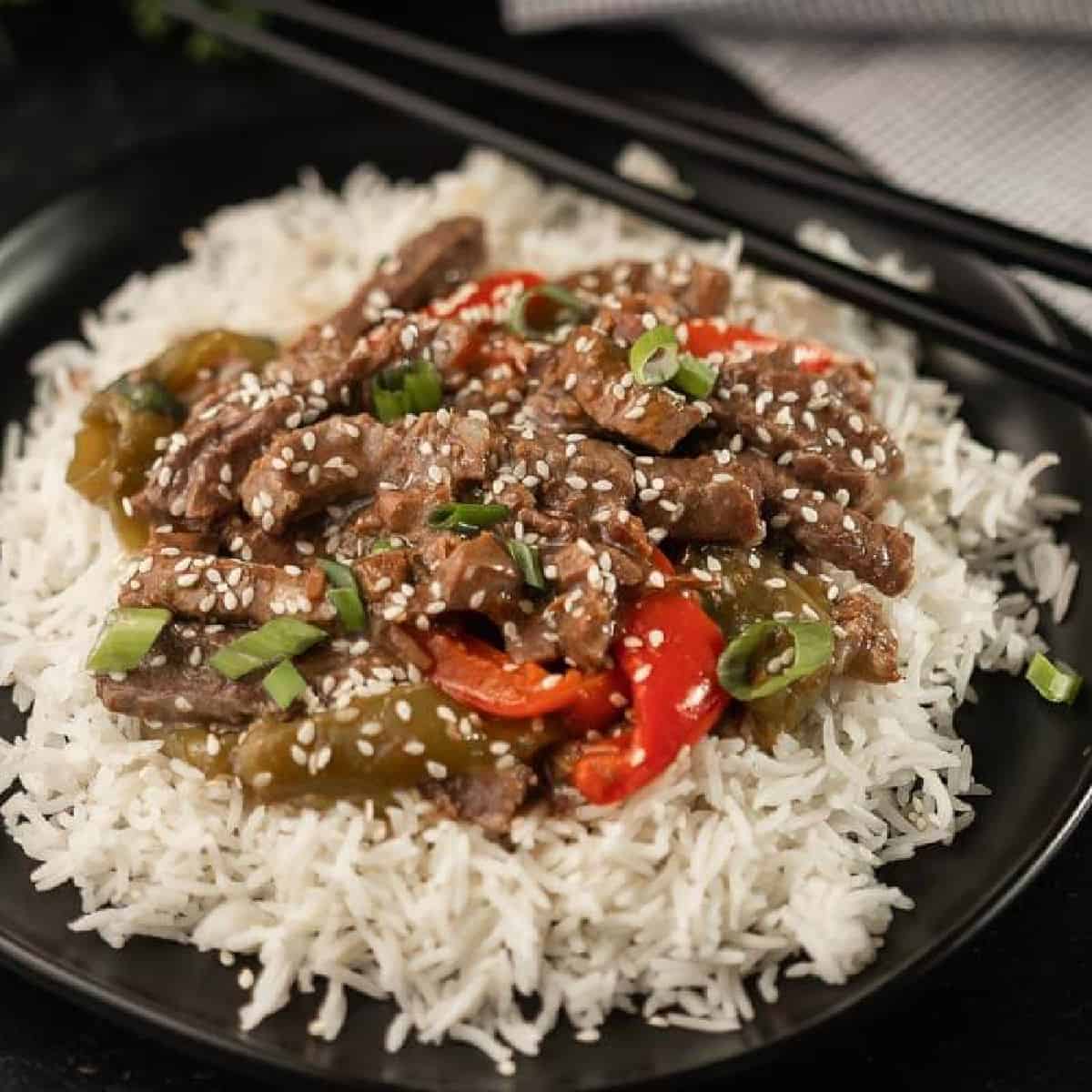 A plate of white rice topped with stir-fried beef strips, red and green bell peppers, sliced green onions, and sprinkled with sesame seeds—just like a delicious Crockpot Pepper Steak Recipe. Chopsticks rest in the background.