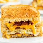 A close-up of a Bacon Grilled Cheese sandwich, with crispy bacon and melted cheddar oozing out between two golden, toasted slices of bread, stacked on a white plate.