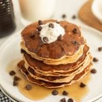 A stack of chocolate chip pancakes topped with whipped cream sits on a white plate, surrounded by syrup and extra chocolate chips. A glass of milk is in the background, making this a perfect chocolate chip pancake breakfast.