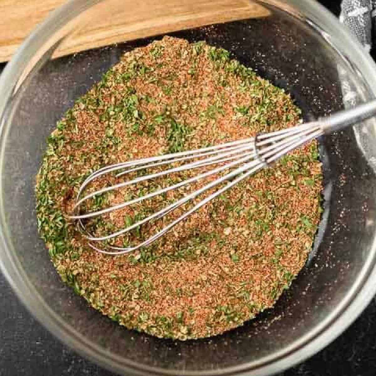 A metal whisk rests in a clear glass bowl filled with a mixed blend of dried spices and herbs, perfect for a homemade Blackened Seasoning Recipe, sitting on a dark countertop.