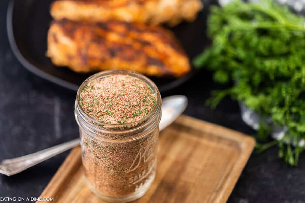 Forget buying blackened spices when you can make this easy Homemade Blackened Seasoning Recipe at home for a lot less. Enjoy Blackened fish, chicken or shrimp! You’ll love this easy cajun spices recipe. It really is the best! #eatingonadime #homemadeseasoningmixes #blackenedseasoningmix A glass jar filled with a Blackened Seasoning Recipe sits on a wooden tray with a spoon beside it. Blurred in the background are grilled chicken pieces on a black plate and a bunch of fresh herbs.