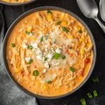 A bowl of creamy Instant Pot Buffalo Chicken Chili topped with shredded cheese, blue cheese crumbles, and sliced green onions. The soup features shredded chicken, corn, and diced tomatoes. Two spoons and fresh green onions are nearby.