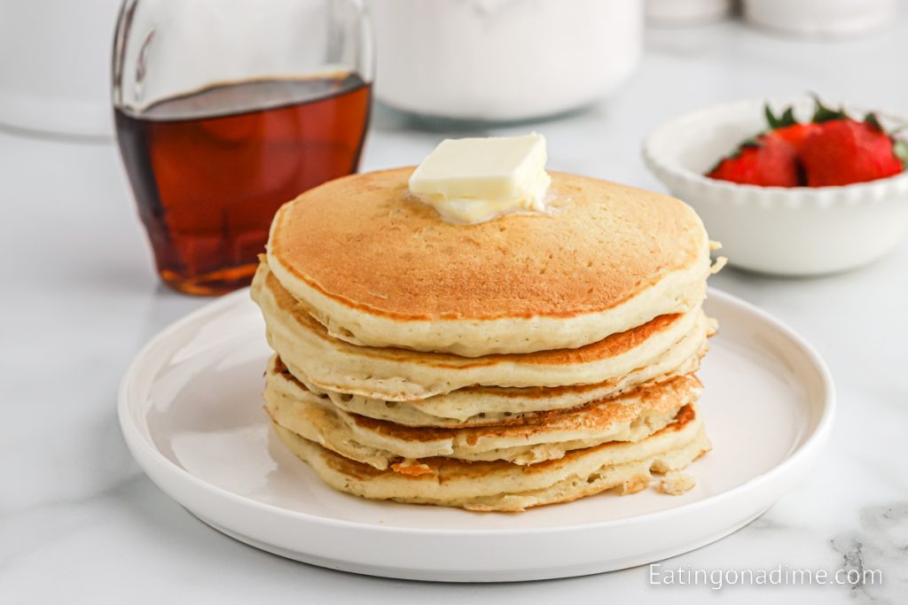 Stack of pancakes on a plate with syrup and a bowl of fresh fruit