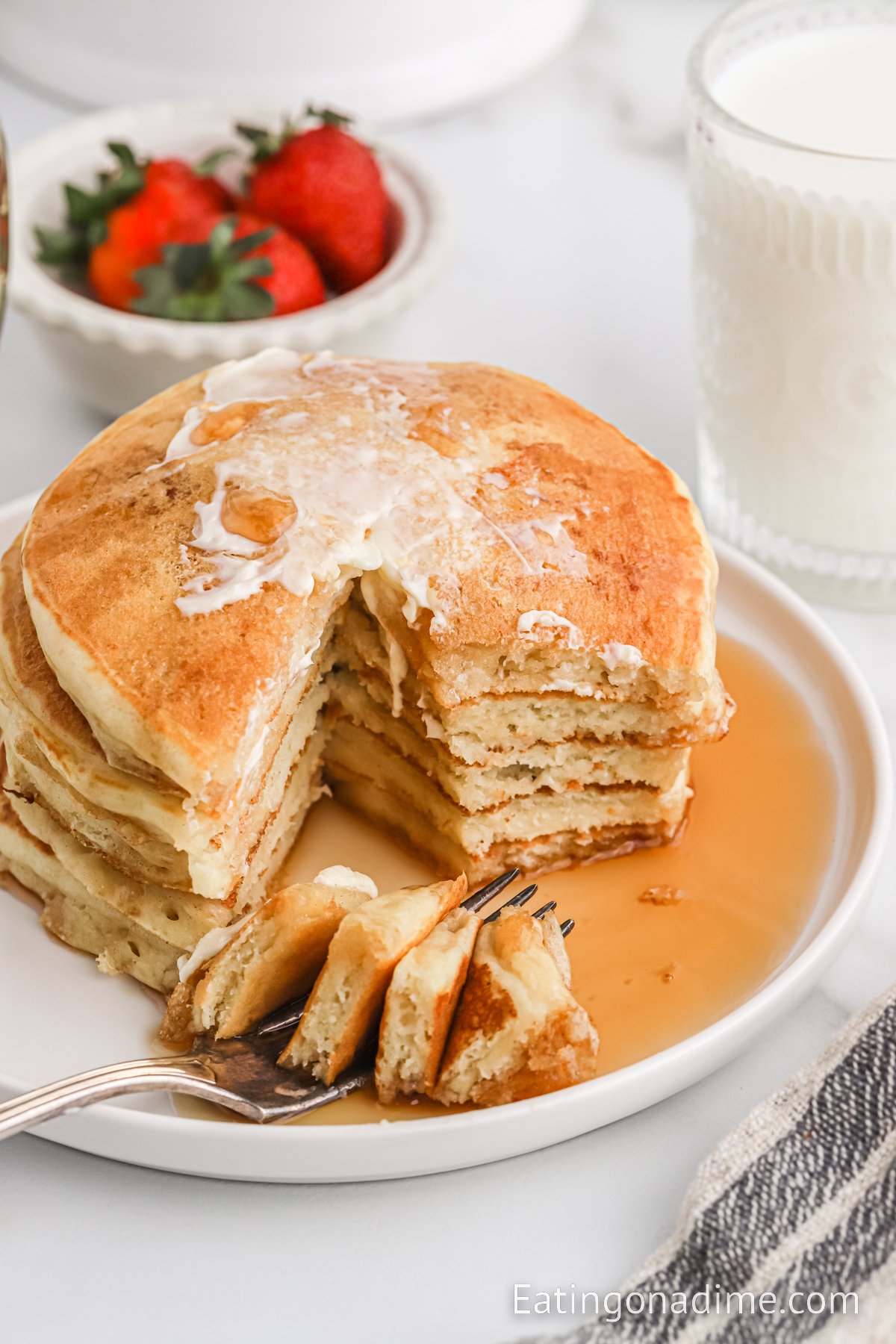 Stack of pancakes on a plate with syrup and a bowl of fresh fruit