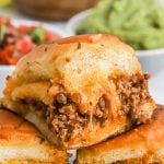Close-up of a cheesy Taco Slider with seasoned ground beef filling on a soft bun, with bowls of guacamole and salsa blurred in the background.