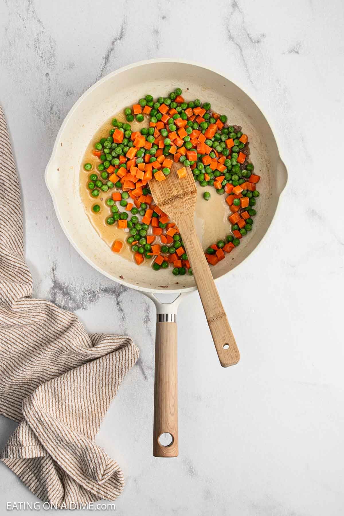 Peas and and diced carrots in a large skillet with a wooden spoon and cooking in oil.