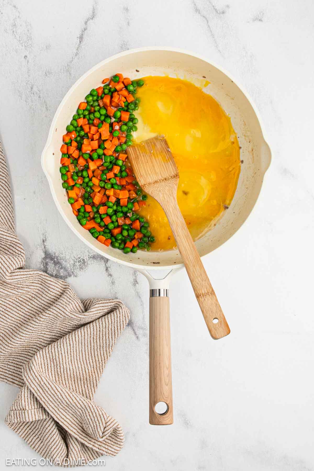 Peas and carrots heating in a skillet with eggs and a wooden spoon.