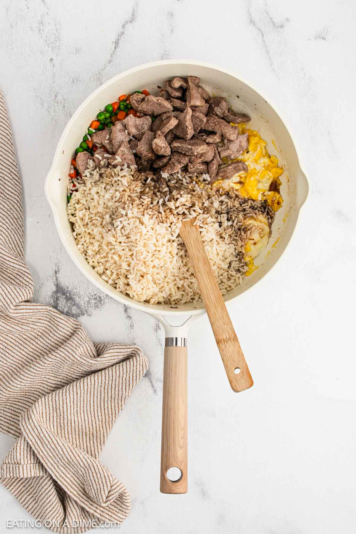 Rice, cooked pork, peas and carrots and eggs cooking a skillet with a wooden spoon.