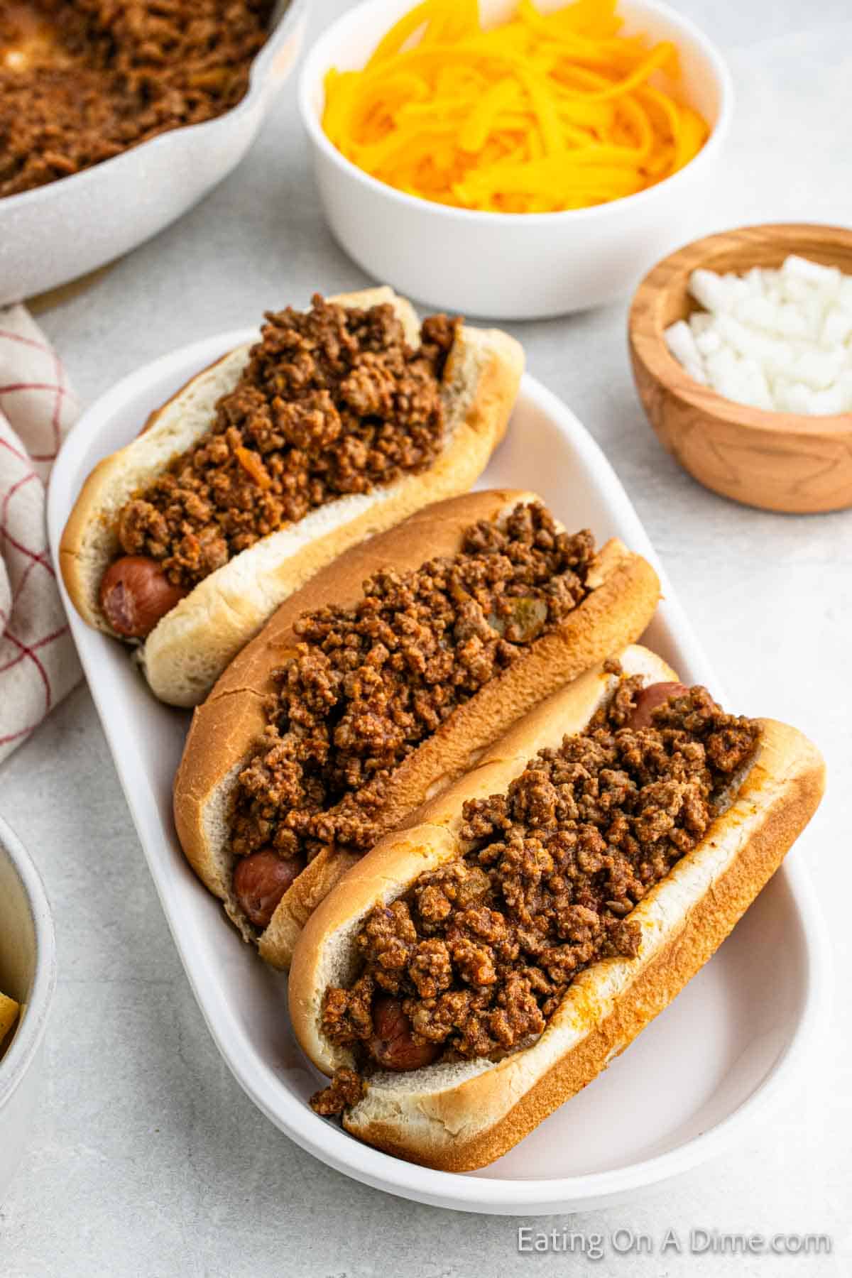 Hot dogs topped with chili on a platter with a bowl of diced onions on this side