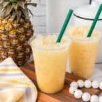 Two plastic cups filled with a yellow, icy pineapple drink sit on a wooden board. Reminiscent of what's in the Paradise Drink at Starbucks, the scene features a fresh pineapple, striped towel, white beads, and green straws for a bright, refreshing vibe.