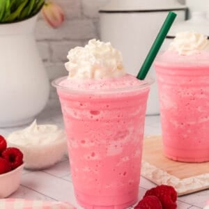 A pink Cotton Candy Frappuccino topped with whipped cream and a green straw sits on a counter next to a bowl of raspberries, with another similar drink in the background. The scene is bright and inviting.