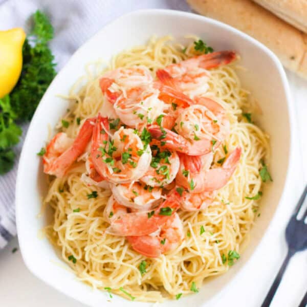 A white bowl filled with pasta topped with shrimp and chopped parsley, placed on a light surface. A lemon and some fresh herbs are partially visible in the background, suggesting a fresh and appetizing meal, related to a crock pot shrimp scampi recipe.