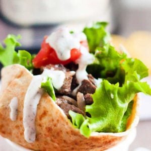 A pita filled with chunks of beef, fresh lettuce, diced tomatoes, and drizzled with a creamy sauce. The pita is presented on a white plate against a blurred background, emphasizing the vibrant colors of the ingredients.