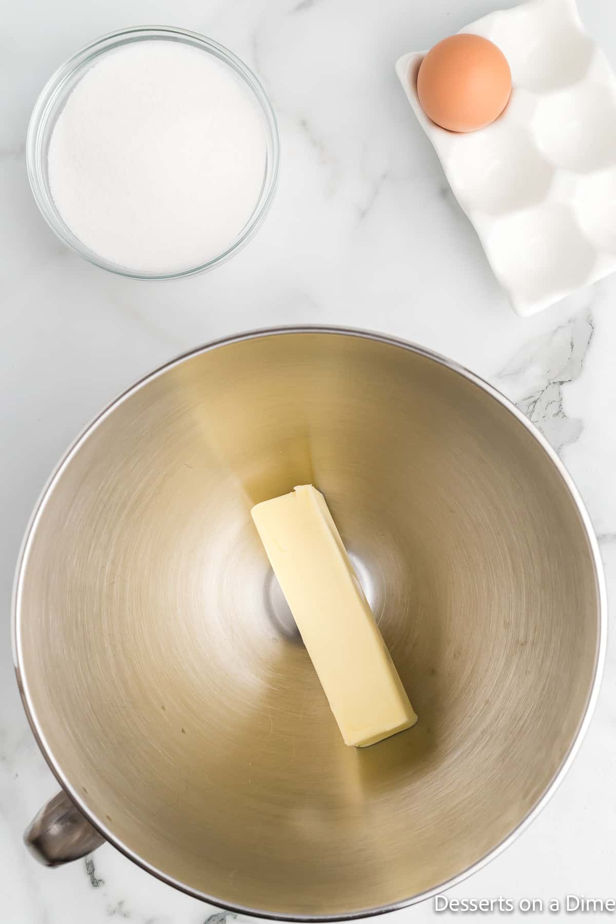 Stick of butter in a bowl with a bowl of sugar and one egg in a carton