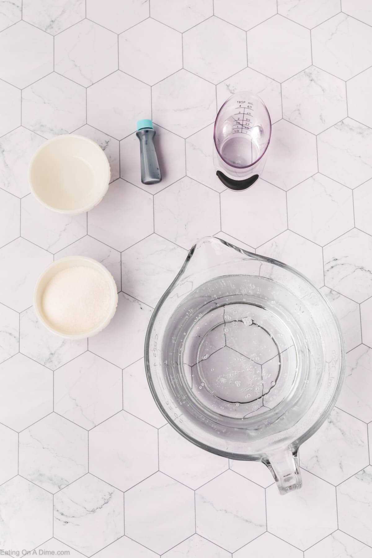 Top view of a marble-patterned surface showcasing the essentials for an easy sonic ocean water recipe: a large measuring cup filled with water, a smaller measuring cup, two bowls with white contents, and a small bottle of blue liquid. They are all arranged neatly on a hexagonal tile pattern.