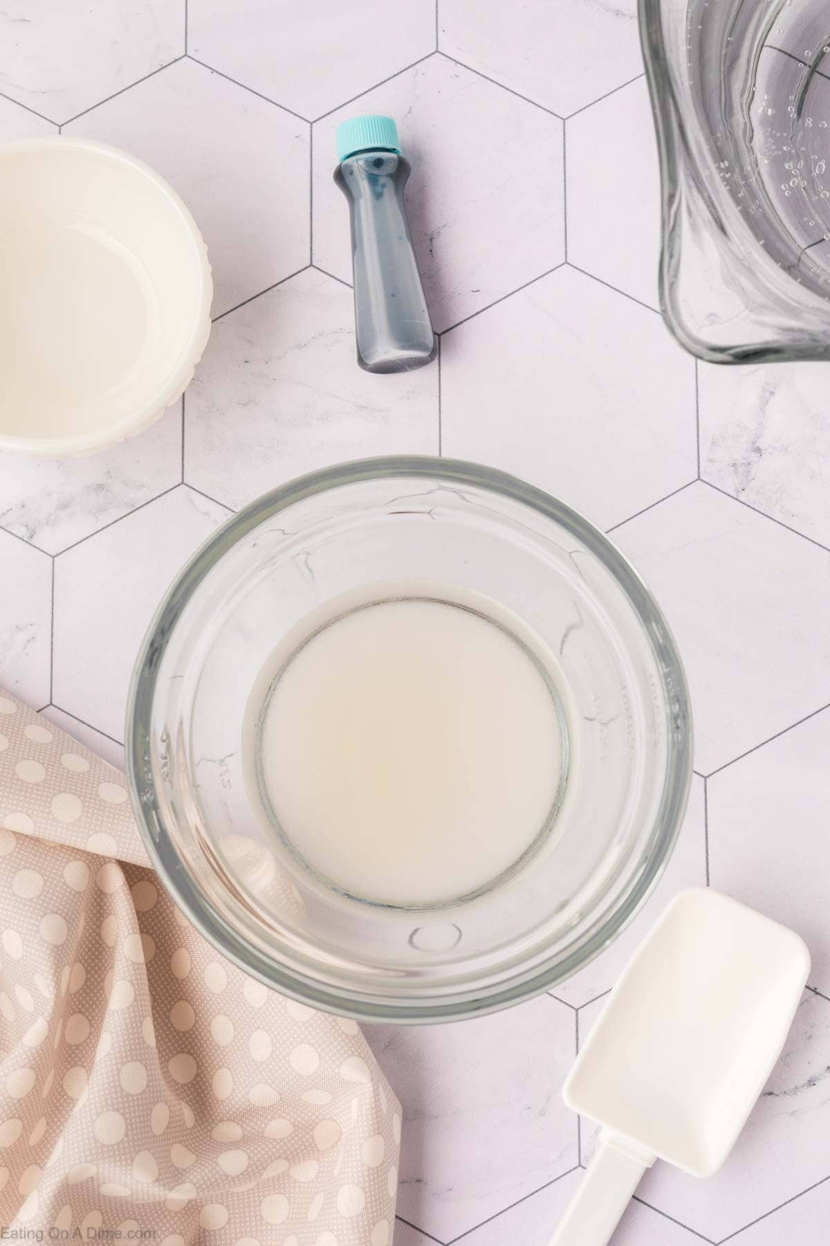 A glass bowl with a white liquid sits on a hexagonal patterned surface, evoking the calm of an easy sonic ocean water recipe, surrounded by a polka dot cloth, a white spatula, a small blue bottle, and another container.