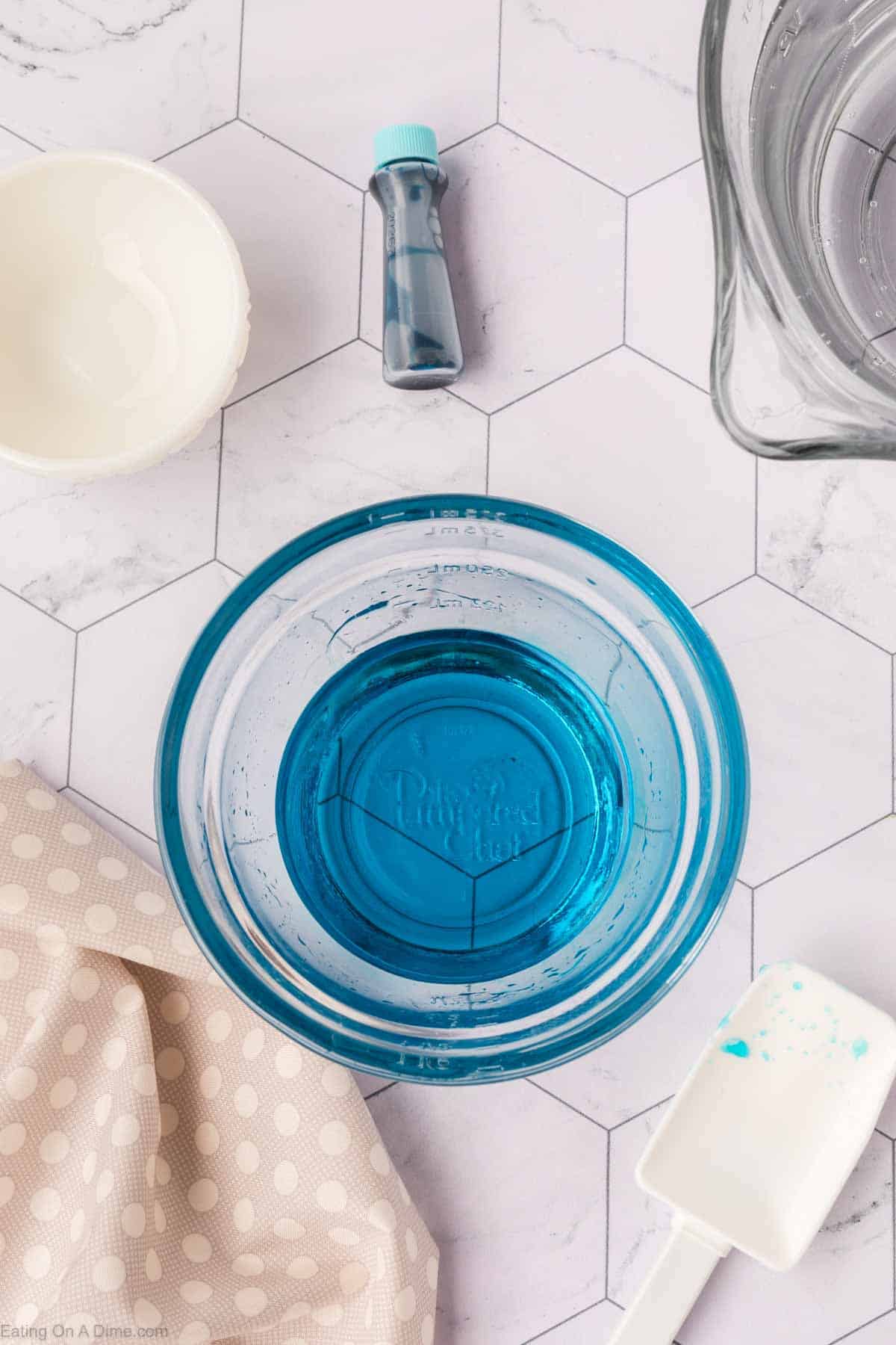 An overhead view reveals a hexagon-patterned marble surface. A glass bowl brims with blue liquid, evoking an easy Sonic ocean water recipe. Nearby are an empty white bowl, a white spatula with a blue stain, a small blue bottle, and a polka-dotted cloth.