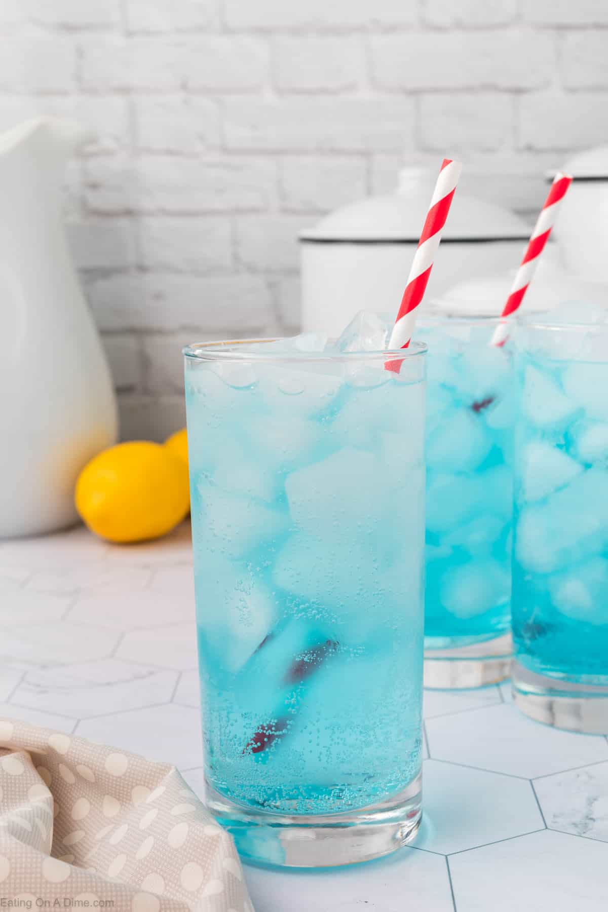 Two tall glasses of blue liquid, resembling an easy Sonic Ocean Water recipe, with ice cubes and red striped straws. The glasses sit on a white hexagonal tile counter, accompanied by a lemon, a white jug, and a beige kitchen towel in the background.