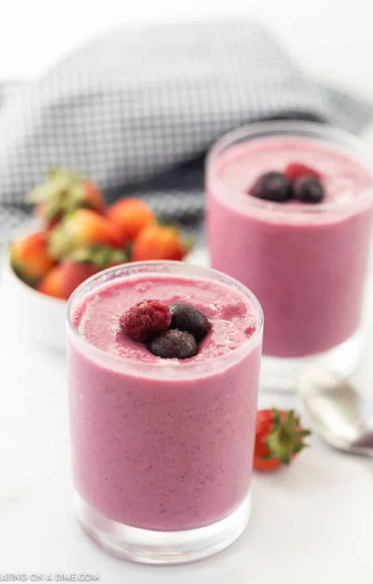 Try this Easy Frozen Fruit Smoothie Recipe for a delicious snack or quick meal. It is such an Easy Fruit Smoothie Recipe.It's also a healthy smoothie recipe. Even the kids will love this Easy smoothie recipe. Try it with strawberries, blueberries and more! Two glasses of pink berry smoothie topped with mixed berries, with a bowl of strawberries and a checkered cloth in the background. An easy fruit smoothie recipe shines here, with a spoon and strawberry on the white surface nearby.