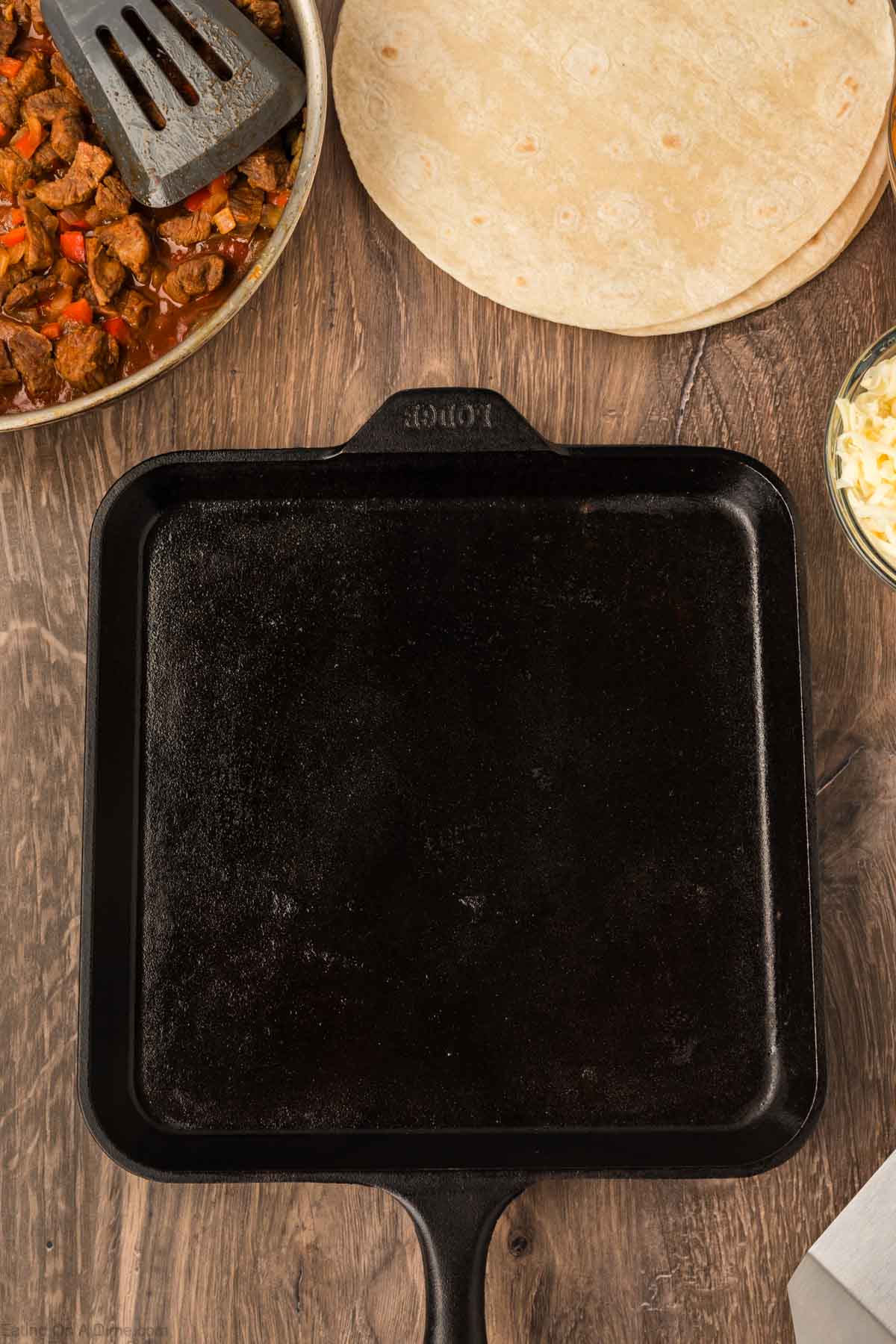 A square, black cast iron skillet sits on a wooden table, surrounded by flour tortillas, a metal spatula in a pan with cooked meat and vegetables for Steak Fajita Quesadillas, and a bowl of shredded cheese.
