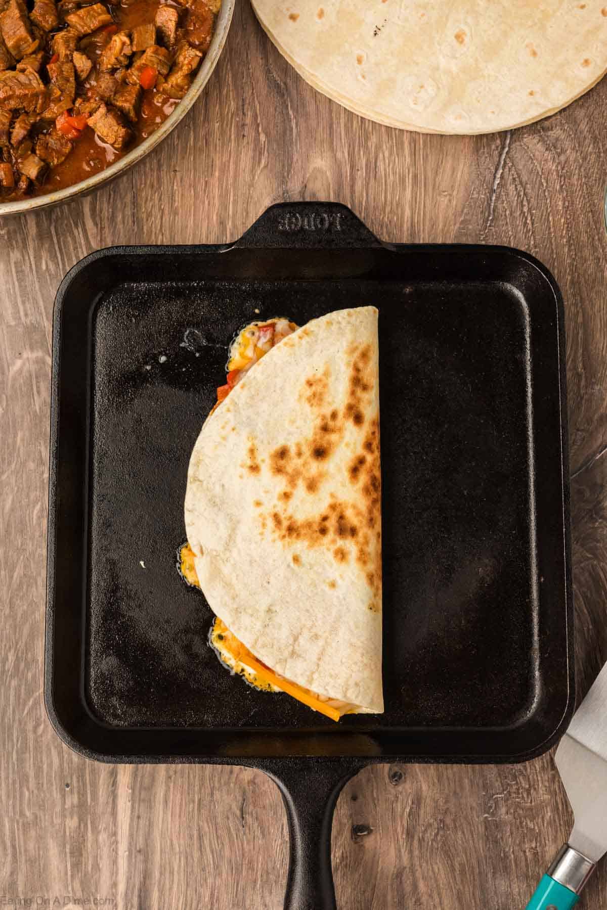 A quesadilla cooking on a black square skillet, filled with melted cheese and vegetables. Nearby are beef pieces for Steak Fajita Quesadillas, a stack of tortillas, and a metal spatula resting on a wooden surface.