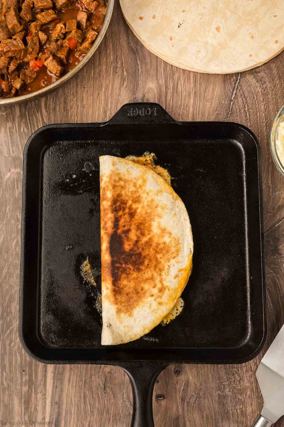 A half-cooked Steak Fajita Quesadilla with melted cheese sizzles on a black skillet. Nearby, a pan of cooked beef and a plain tortilla rest on a wooden surface.