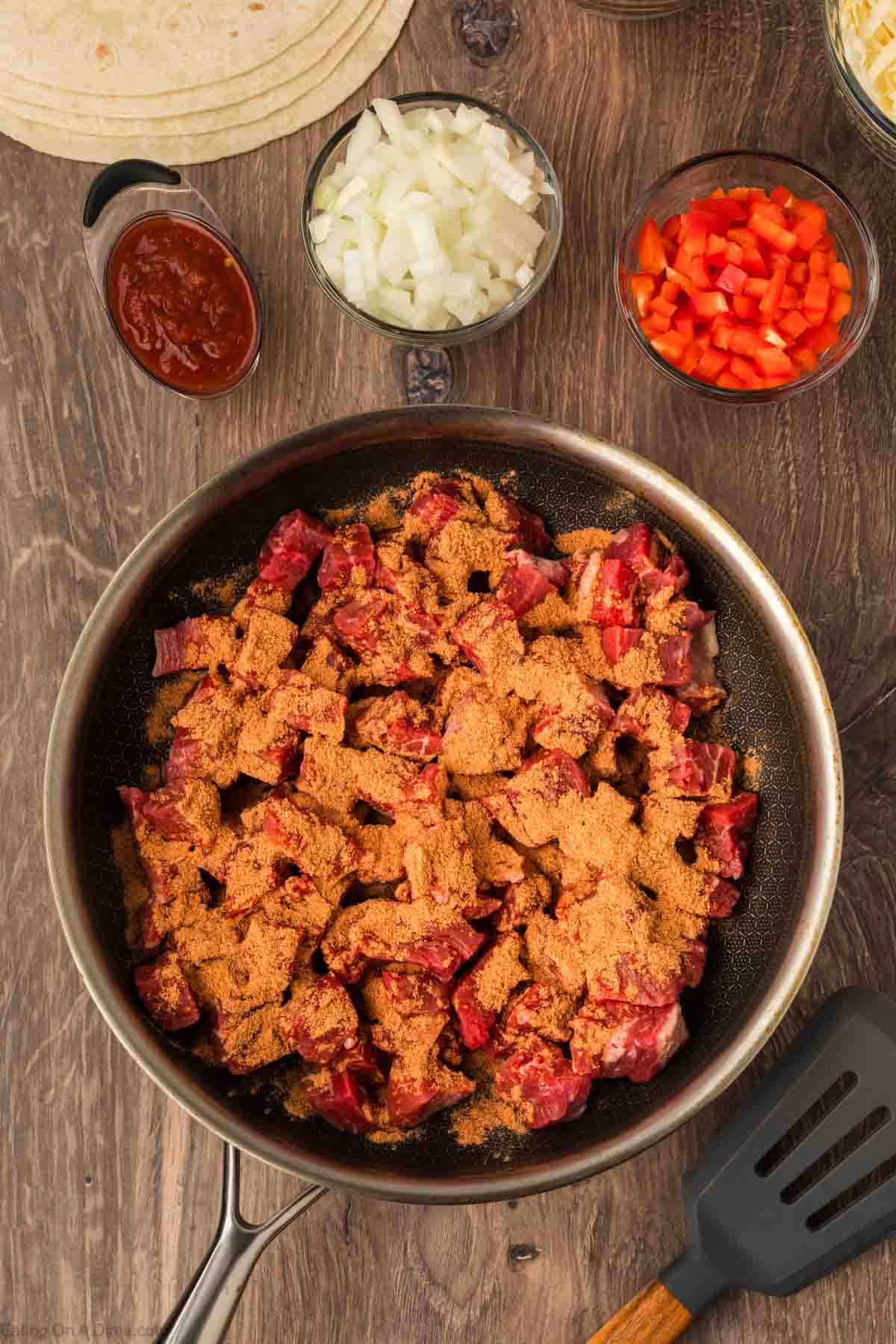 A skillet with raw beef cubes for Steak Fajita Quesadillas topped with taco seasoning, surrounded by bowls of chopped onions, red bell peppers, sauce, shredded cheese, and tortillas on a wooden surface. A spatula rests beside the skillet.