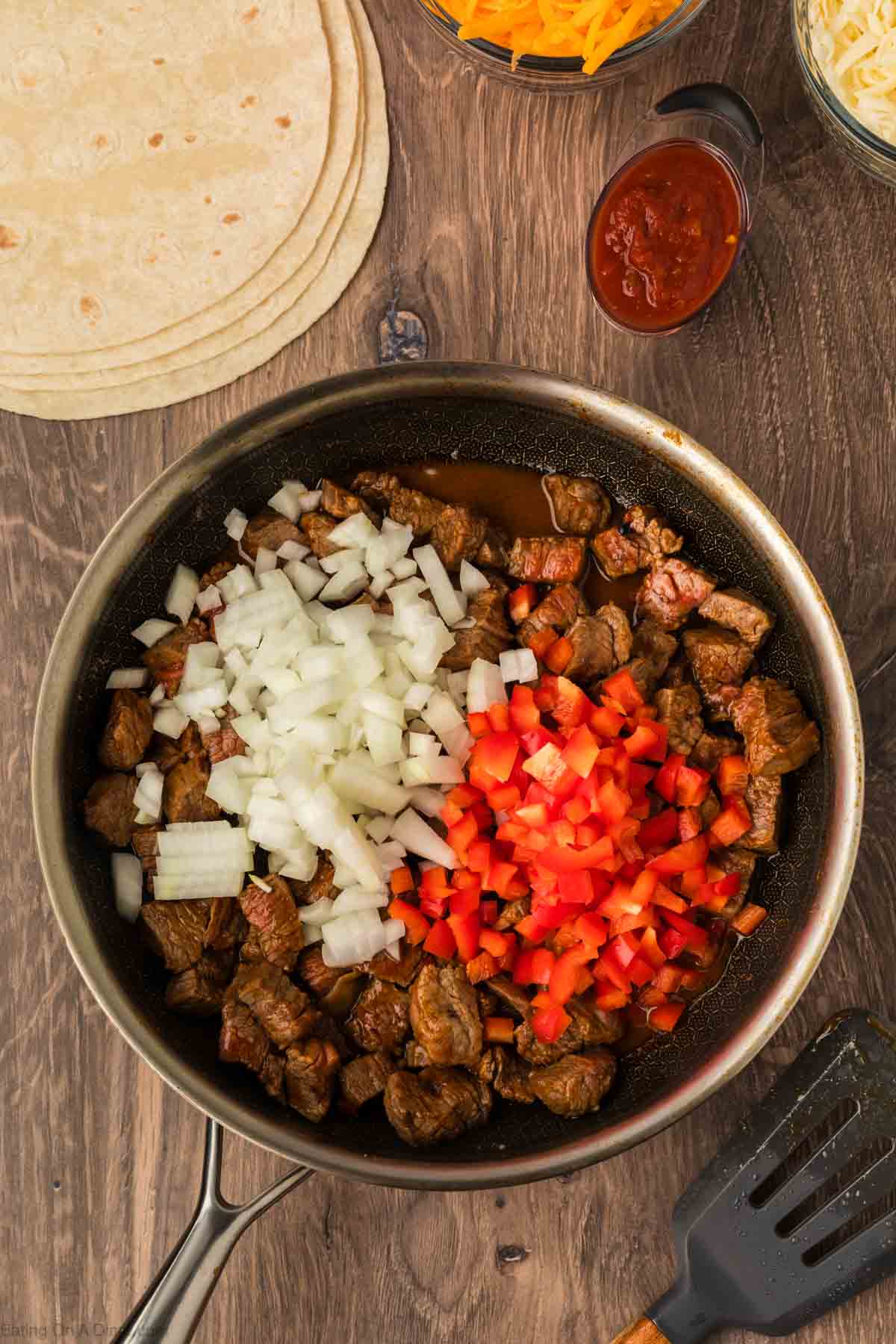 A skillet with cooked beef, chopped onions, and diced red bell peppers on a wooden table, surrounded by flour tortillas, shredded cheese, salsa, and a spatula—perfect ingredients for delicious Steak Fajita Quesadillas.