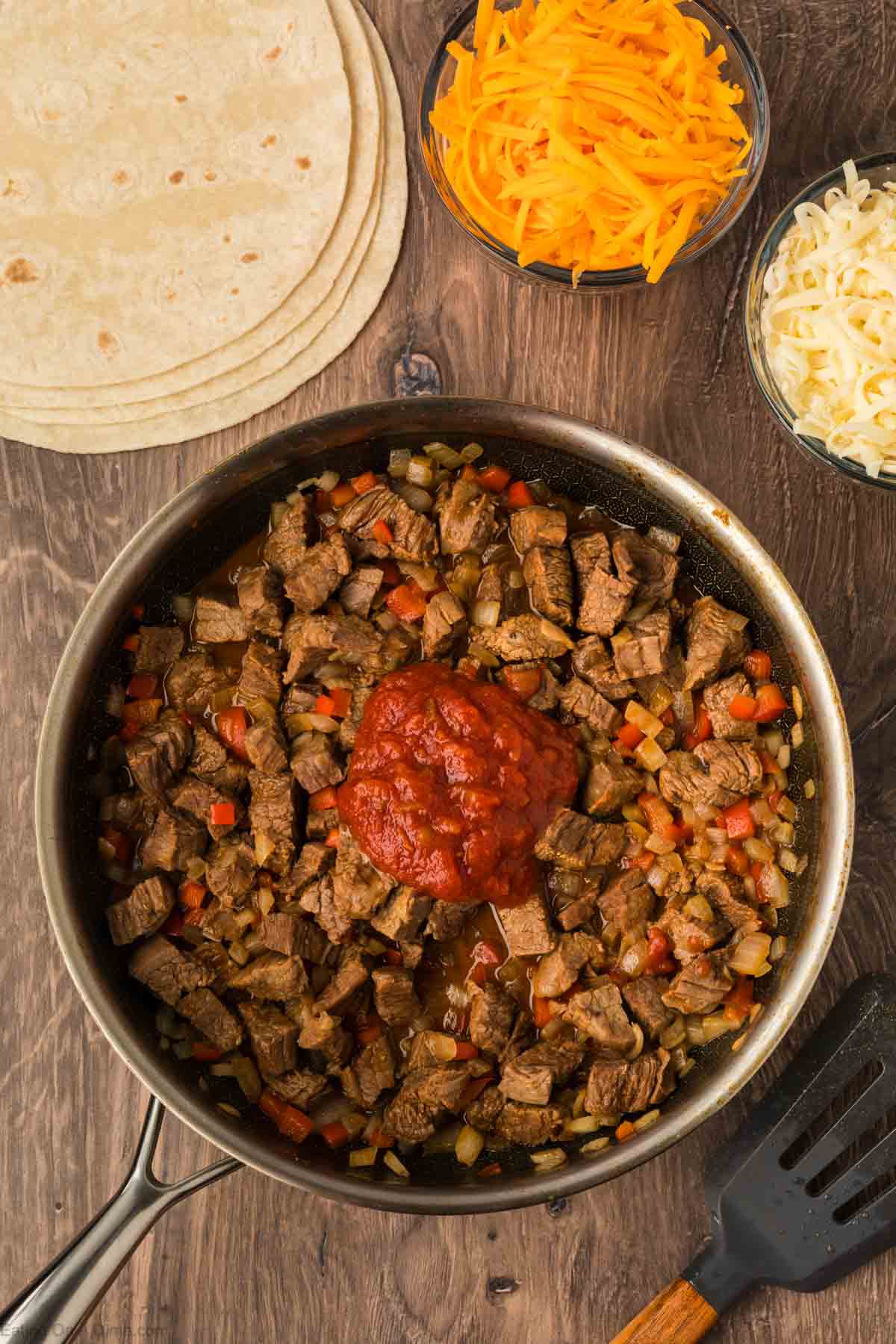 A skillet filled with diced beef, onions, and peppers—perfect for Steak Fajita Quesadillas—sits on a wooden surface. Nearby are shredded cheddar, mozzarella, tortillas, and a scoop of salsa in the center.