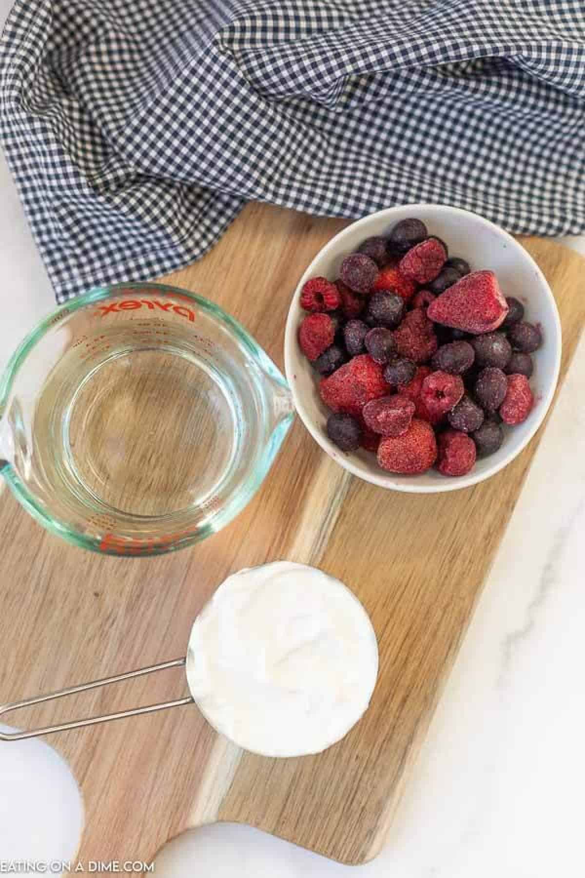 A wooden board holds the ingredients for an easy fruit smoothie recipe: a bowl of frozen mixed berries, a measuring cup of water, and a cup of yogurt. A checkered cloth is partially visible at the top of the image.