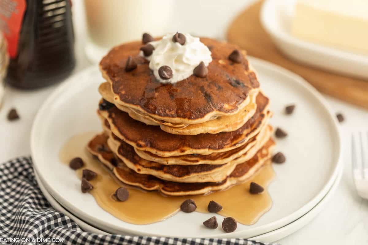 A stack of chocolate chip pancakes topped with whipped cream sits on a white plate, surrounded by syrup and extra chocolate chips. A glass of milk is in the background, making this a perfect chocolate chip pancake breakfast.