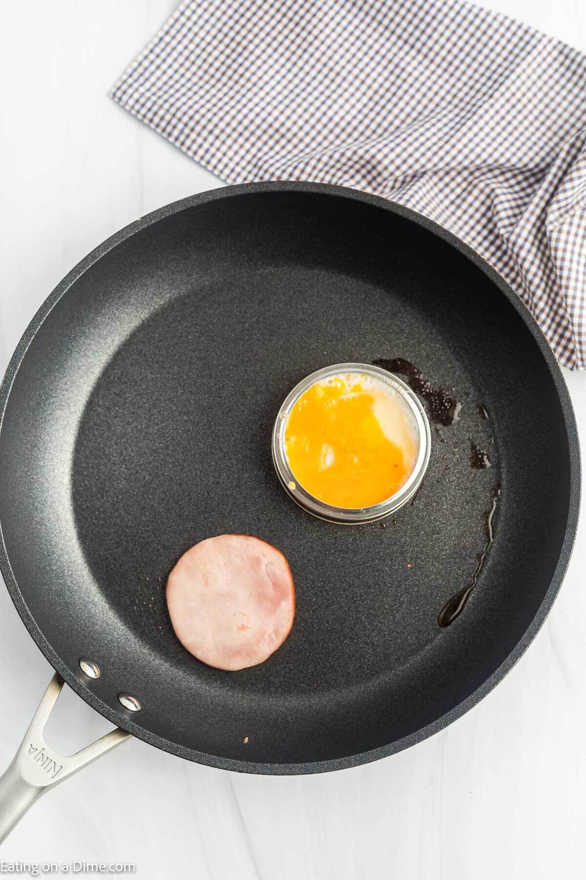 Canadian bacon and a cracked egg in a mason jar lid in a skillet with melted butter.
