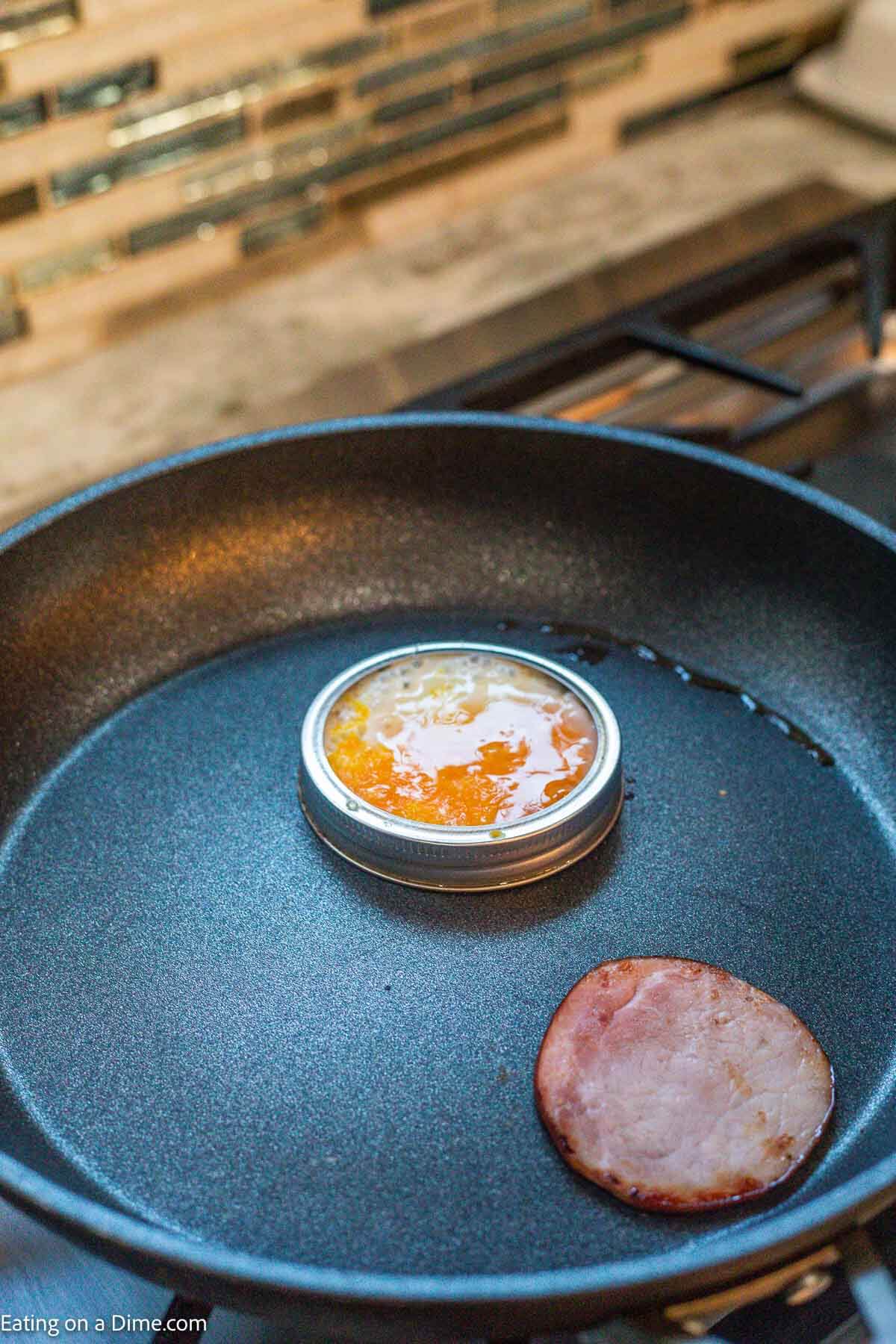 Cooked canadian bacon slice and egg cooking in a mason jar lid in a skillet.
