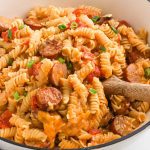 A close-up of creamy Kielbasa Pasta with rotini, sliced sausage, diced tomatoes, and chopped green onions, served in a white bowl with a wooden spoon.