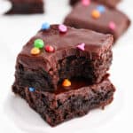 Two thick, fudgy brownies stacked on a white surface, topped with glossy chocolate frosting and colorful round sprinkles—a homemade take on the Cosmic Brownies recipe. The top brownie has a bite out of it, revealing its irresistibly moist texture.