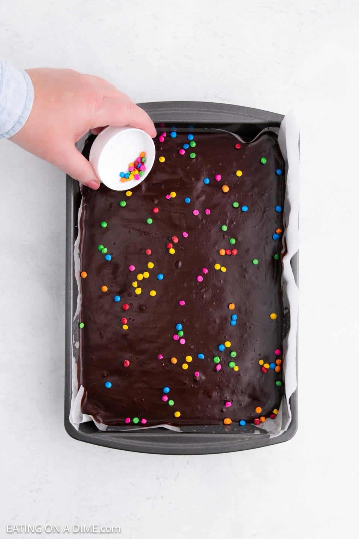 Adding sprinkles over the top of the brownies in a baking dish lined with parchment paper