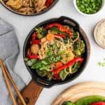 A black skillet filled with noodle stir fry inspired by a Crock Pot Chicken Lo Mein Recipe, featuring snow peas, broccoli, carrots, red bell peppers, and sesame seeds, sits on a wooden board beside a bowl of green onions and chopsticks.