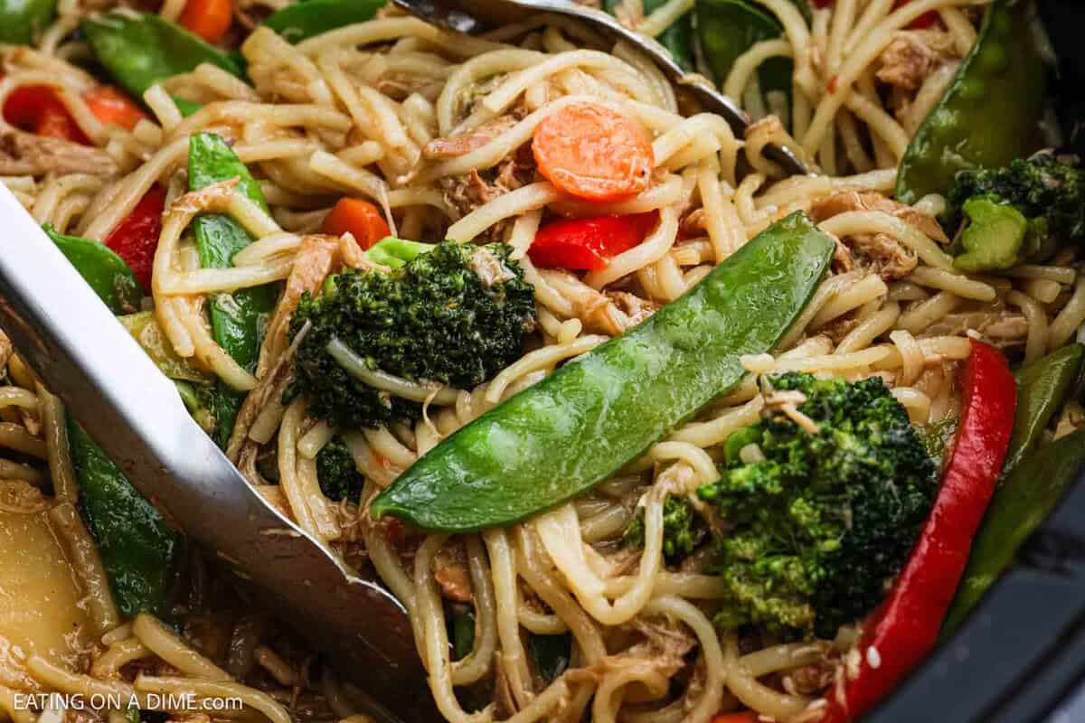 Close-up of Crock Pot Chicken Lo Mein Recipe: stir-fried noodles with broccoli, snap peas, carrots, and red bell peppers, mixed with sauce and shredded meat, being served with metal tongs.
