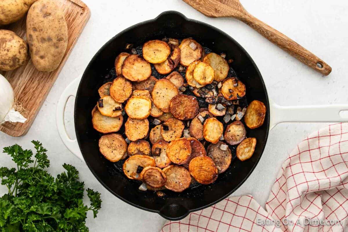 A skillet of pan-fried potatoes with crispy edges and sautéed onions sits on a white surface. Beside it are whole potatoes, a wooden spoon, a checkered napkin, and sprigs of fresh parsley.