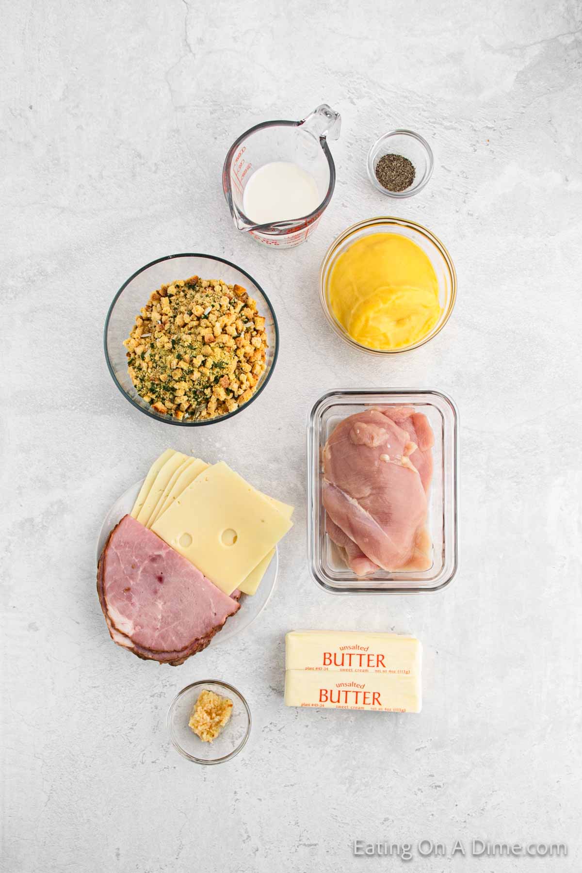 Minced garlic in a bowl, sticks of butter a plate of slice ham and swiss cheeese, chicken breast, stuffing, cream of chicken soup, milk, pepper