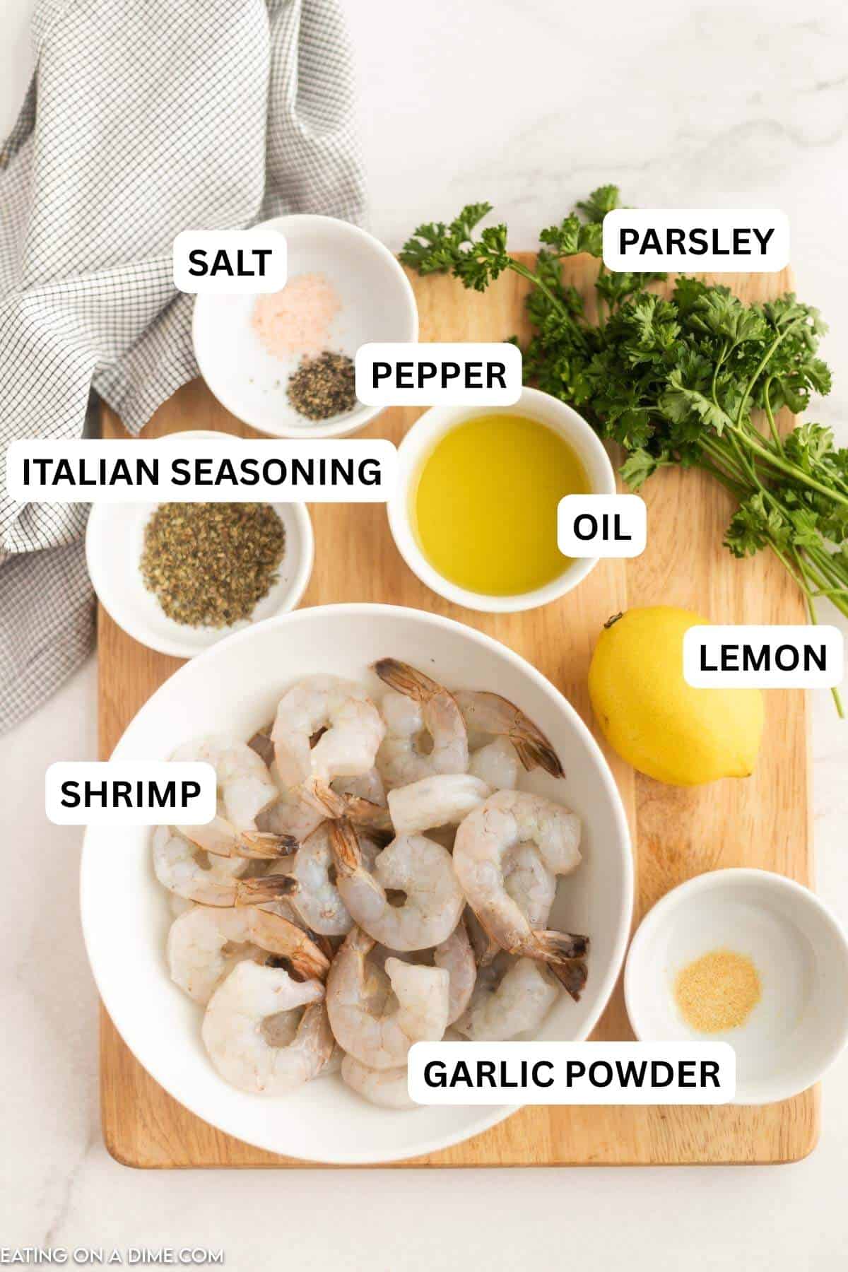 A white bowl of raw peeled shrimp on a wooden cutting board with olive oil, fresh parsley, a lemon, and small bowls of spices.